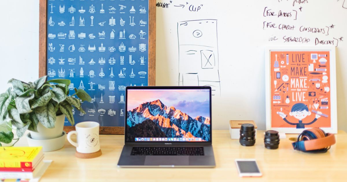 A workspace with a laptop, plant, books, framed print, headphones, and camera lenses on a desk. Whiteboard in the background.