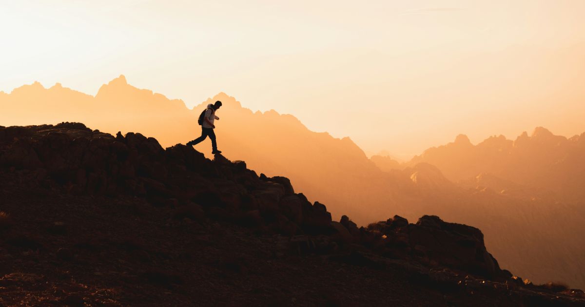 Silhouette of a person hiking uphill on a rocky terrain during sunrise or sunset with rugged mountains in the background.